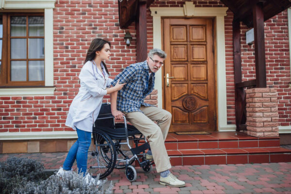 A healthcare provider in a white coat with a stethoscope assists an elderly man as he transfers from a wheelchair onto the front steps of a brick home.