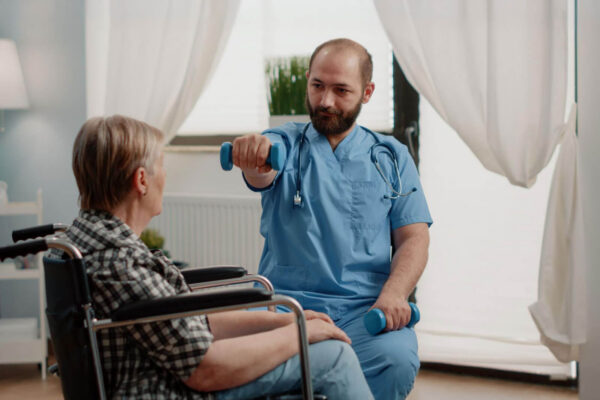 A home healthcare professional demonstrates a dumbbell exercise for a patient seated in a wheelchair.
